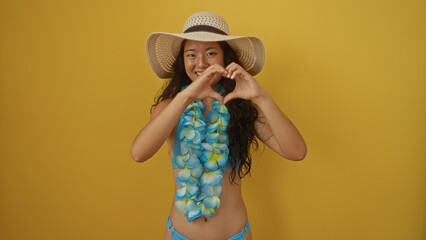 Beautiful young chinese woman in a blue swimsuit and flowers lei making a heart gesture with hands over isolated yellow background
