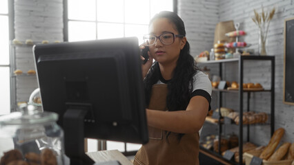 Young chinese woman working in a bakery shop, operating a computer and answering the phone, with...