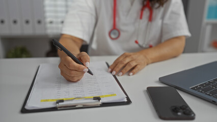 Woman writing notes with pen in clinic office showing mature female in medical setting using laptop and clipboard, highlighting professional hands in hospital environment.