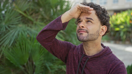 Handsome young hispanic man looks up in urban park setting surrounded by greenery under bright sunlight