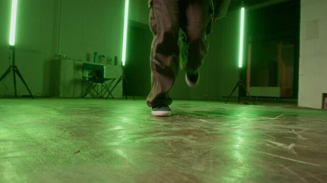 Cropped slowmo shot of legs of anonymous man in urbanwear practicing top rock movements of breakdance in room with green led lamps