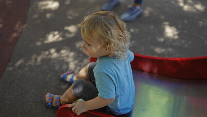 Naklejka premium Toddler boy with blonde hair playing on a red slide in an urban park on a sunny day