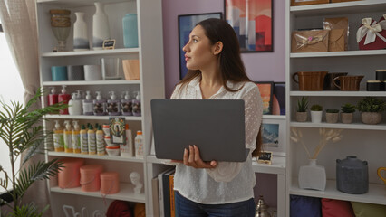 Young woman holding laptop in home decor store, surrounded by various decorative items and plants, looking away thoughtfully