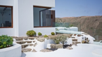 Modern house exterior with blurred white walls, outdoor steps, and surrounding greenery in a sunny setting with a blurred background view of hills and clear sky.