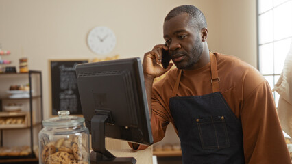 Young man working in a bakery, talking on the phone while facing a computer, surrounded by baked goods in an indoor shop setting