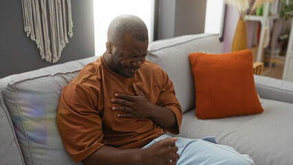 Young man suffering heart pain while sitting in the living room at home, clutching his chest with a painful expression