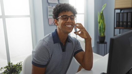 Young man talking on phone in an office, smiling while seated at his desk, wearing glasses and a casual shirt, with documents and a potted plant visible in the background
