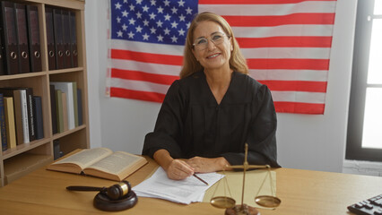 Mature woman judge sitting at a desk in an office courtroom with an american flag in the...