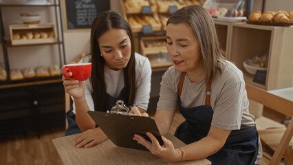 Two women bakers discussing work while one holds a red cup and the other writes on a clipboard in a cozy bakery shop.