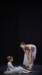 Adult ballerina bending to adjust pointe shoes, child sitting nearby holding ribbons, both in white...