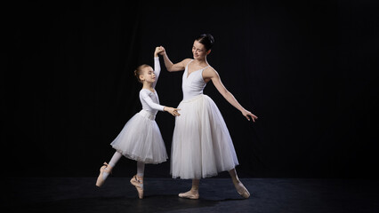 Fototapeta premium Adult ballerina assisting child in dance position, both smiling gently while holding hands, dressed in white ballet costumes, against black background. Concept of ballet, elegance and beauty, grace.
