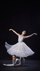 Fototapeta premium Adult ballerina spinning elegantly in tutu while young ballerina poses on floor thoughtfully both dressed in white, against black studio background. Concept of ballet, elegance and beauty, grace. Ad