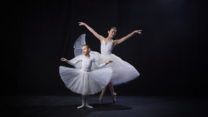 Naklejka premium Little kid and ballerina in white tutus gracefully perform synchronized arm movements, displaying elegance and poise against black background. Concept of ballet, elegance and beauty, grace. Ad