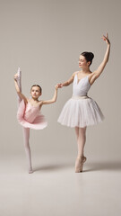 Naklejka premium Ballerina in white tutu supports little girl, dancer in pink tutu performing leg stretch, emphasizing guidance and flexibility against white background. Concept of ballet, elegance and beauty, grace.