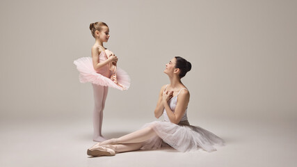 Adult ballerina sits gracefully on floor, watching young girl in pink tutu with admiration as they engage in heartfelt interaction against grey background. Concept of ballet, elegance and beauty. Ad