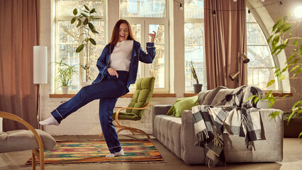 Happy, smiling young woman in stylish blue pajamas cheerfully dancing and playing imaginative guitar in cozy, warm living room at home. Concept of happiness, carefree lifestyle, relaxation