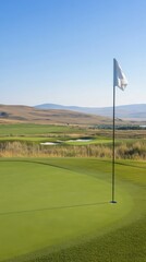 A panoramic view of a serene golf course with rolling hills under a clear blue sky. The lush green field features a flag on the green, adding to the tranquil landscape.