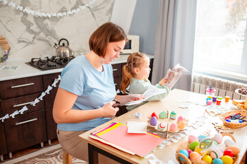 Fototapeta premium Side view of young Caucasian mother and her daughter carve a bunny mask for the Easter spring holiday. The family is sitting at the kitchen table and making festive decorations