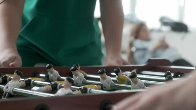 After goal in kicker teenager takes ball out of special hole and puts it into game, static shot. Friends play table football and actively rotate handles with figures of football players.