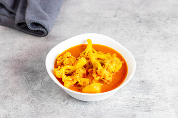 a bowl of vibrant orange-yellow curry with cauliflower florets and potatoes, placed on a Gray textured surface beside a towel.