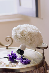 White carnation bridal bouquet with purple flowers and wedding invitation on a vintage table.