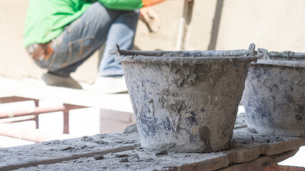 Close-up of two plastic buckets of cement placed on a plank that hangs on scaffolding, and in the background there are workers working on plastering the building's walls.