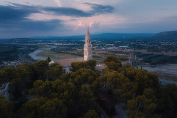 Fototapeta premium Aerial view of a historic church tower in a scenic valley during sunset, surrounded by trees and a meandering river