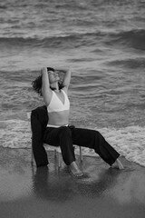 A black-and-white photo of a young Asian woman sitting on a chair in the water, tilting her head...