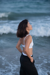 A young Asian woman stands on the beach in white lingerie and black pants, gazing at the waves, with the ocean in the background