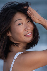 Fototapeta premium close-up portrait of an Asian woman on the beach, gently fixing her hair with her hands, looking into the camera, and smiling. Her natural beauty, freckles, and beauty mark above her lip are highlight
