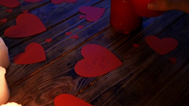 Close up of wooden table with lighted candles and scattered red paper hearts, woman pouring wax from candle on heart shaped card with text I love you.
