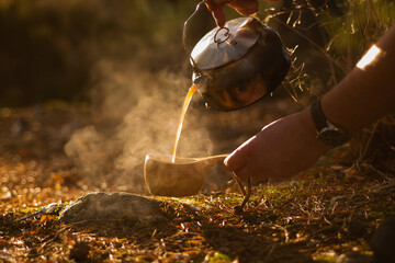 Hands pouring coffee into a wooden cup from a camp kettle in a forest envoronment