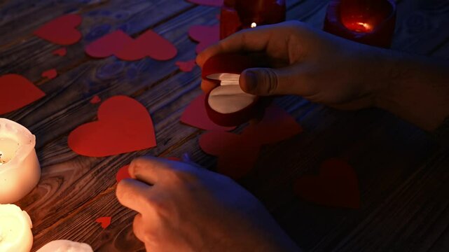 Close up of wooden table in darkness with red and white lighted candles and scattered red paper hearts, man puts diamond ring in the jewelry box and leaves it on the table.