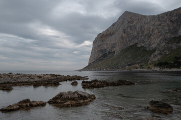 Dramatic coastal landscape along Sicily's Capo Gallo Nature Reserve showcases rugged limestone cliffs meeting turquoise Mediterranean waters. Moody winter skies and scattered rocks 