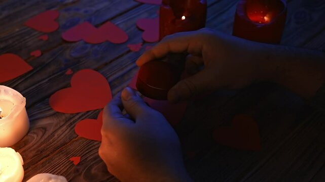 Close up of wooden table in darkness with red and white lighted candles and scattered red paper hearts, man holding and opens jewelry box with diamond ring inside.