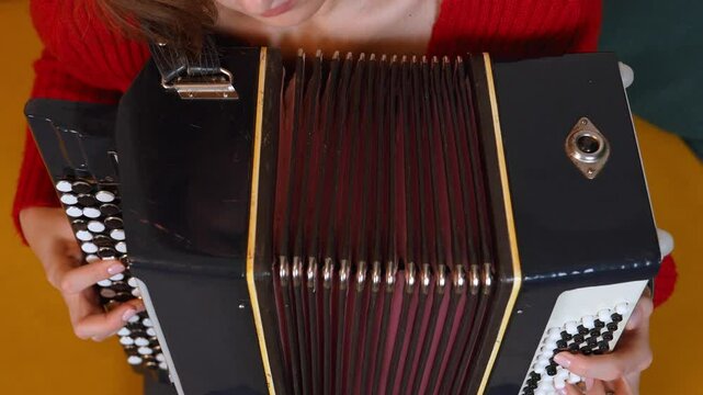 Young female musician in red sweater playing accordion with deeply expressive mimic excitement, capturing musical passion while performing melody with professional technique.