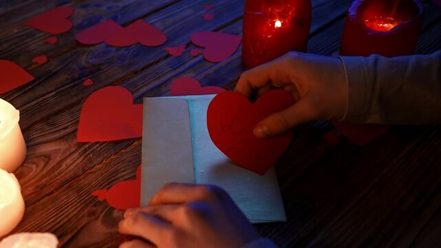 Close up of wooden table in darkness with lighted candles and scattered red paper hearts, woman putting valentines card with text I love you in envelope.