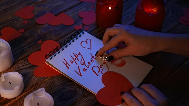 Close up of wooden table with lighted candles and scattered paper hearts, notebook with text happy valentines day laying on the desk, woman puts diamond ring on finger.
