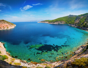 Obraz premium A breathtaking coastal view shows vibrant blue Mediterranean waters meeting rugged cliffs and green hills under a bright blue sky with scattered clouds. 