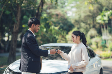 Handing Over the Keys:  A young woman smiles brightly as she receives car keys from a car salesman,...