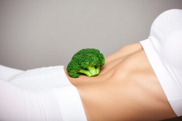 A woman with an athletic gym body and white exercise clothing holds a broccoli on her abdomen in a healthy eating message