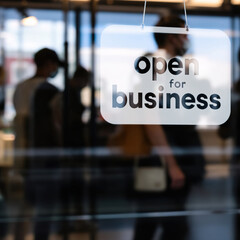 Fototapeta premium A clear open for business sign hangs in a doorway, showcasing a diverse group of people entering a bustling office environment on a bright day, highlighting inclusivity in the workspace