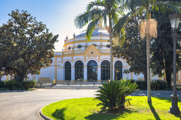Fototapeta premium Seville town, capital of Andalusia in Spain. Elegant rotunda building in a sunny park setting, surrounded by lush greenery and palm trees.