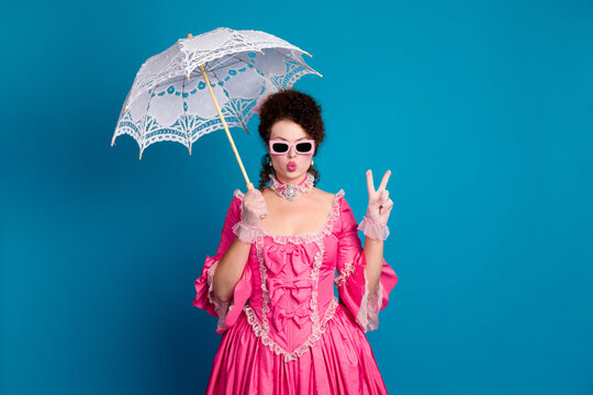 Young woman in pink corset dress with parasol on blue background making peace sign for stylish vintage cosplay