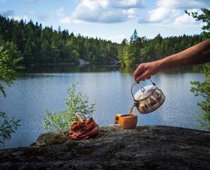 Male hand pouring coffee from a camp kettle into a wooden cup in front of a lake in the forest