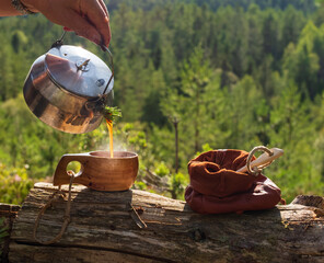 Male hand pouring coffee from a camp kettle into a wooden cup in front of a sush forest