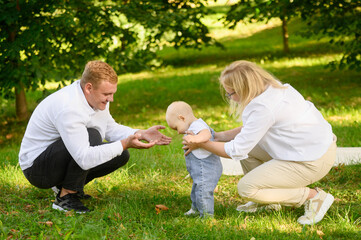 Child walks on grass to meet his dad, holding hands with his mom on grass, toddler learns to walk and takes his first steps. Family concept, family vacation, child's first steps.