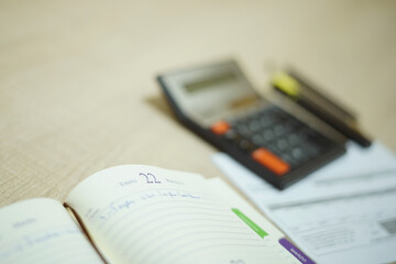 Detalle de una agenda en primer plano a la izquierda en una mesa, al fondo a la derecha papeles, una calculadora y bol&iacute;grafos desenfocado, mesa oficina 