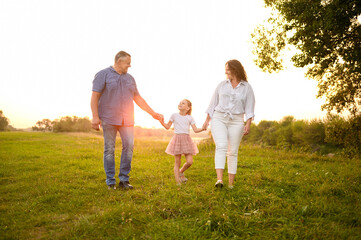 Fototapeta premium Happy family walking together in park at sunset. Mom, dad, daughter spend time together outdoors, walking along path, communicating, spending time together. Happy family, love and parenthood concept.