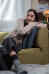 Obraz premium Happy young Asian woman relaxing on a cozy sofa in her living room, enjoying a joyful weekend at home with a serene and comfortable expression